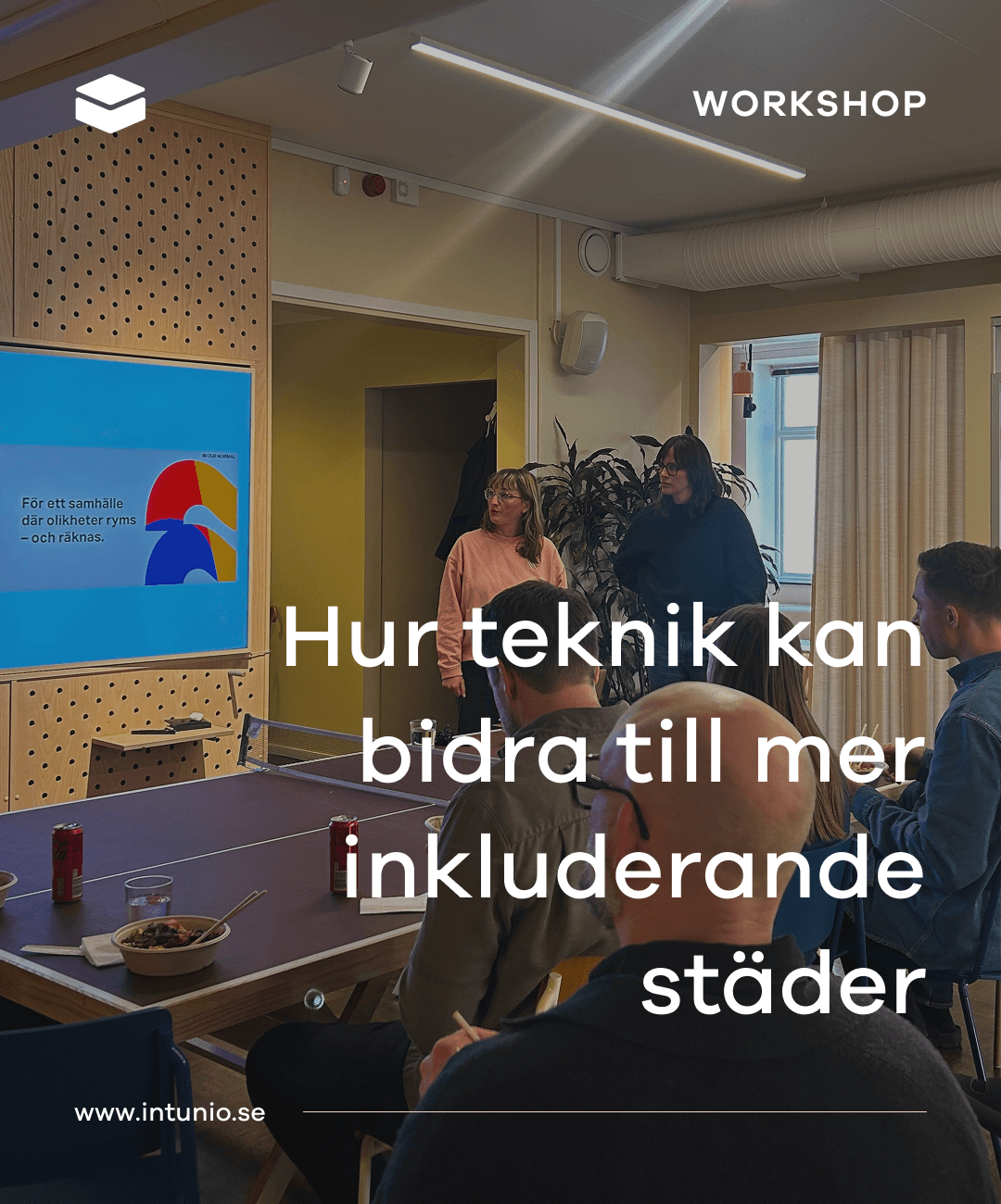 Personer sitter runt ett bord och deltar i en workshop medan två personer presenterar framför en skärm om inkluderande städer.